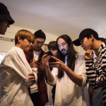 Steve Aoki shows something on his phone to BTS members, who are gathered around him looking at the screen. - midnightrebels.com