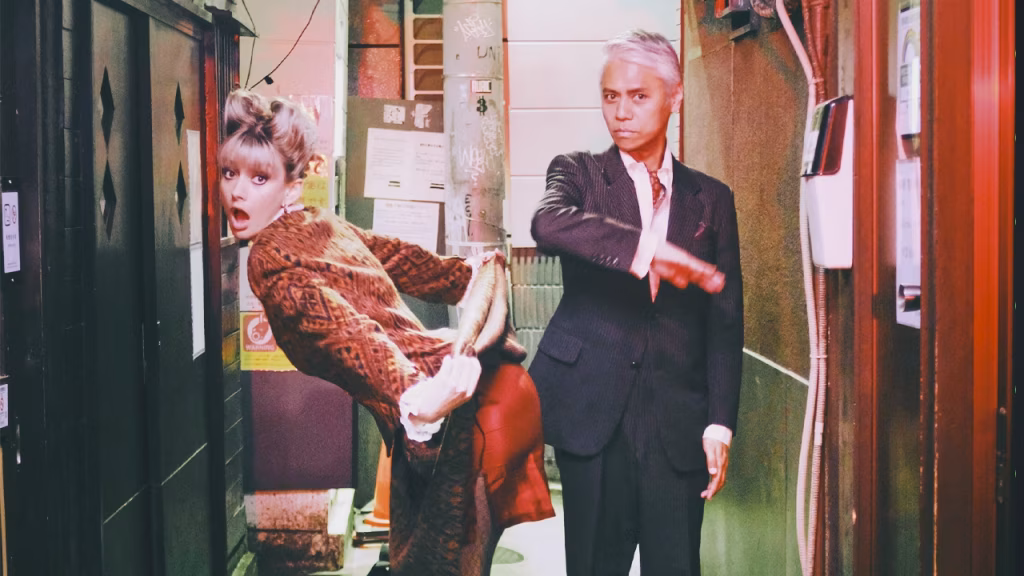 A quirky music video still features a woman in a patterned brown outfit leaning back with a surprised expression and her feet being manipulated, while a man in a pinstripe suit stands beside her with a stern expression in a narrow, urban alleyway. - midnightrebels.com