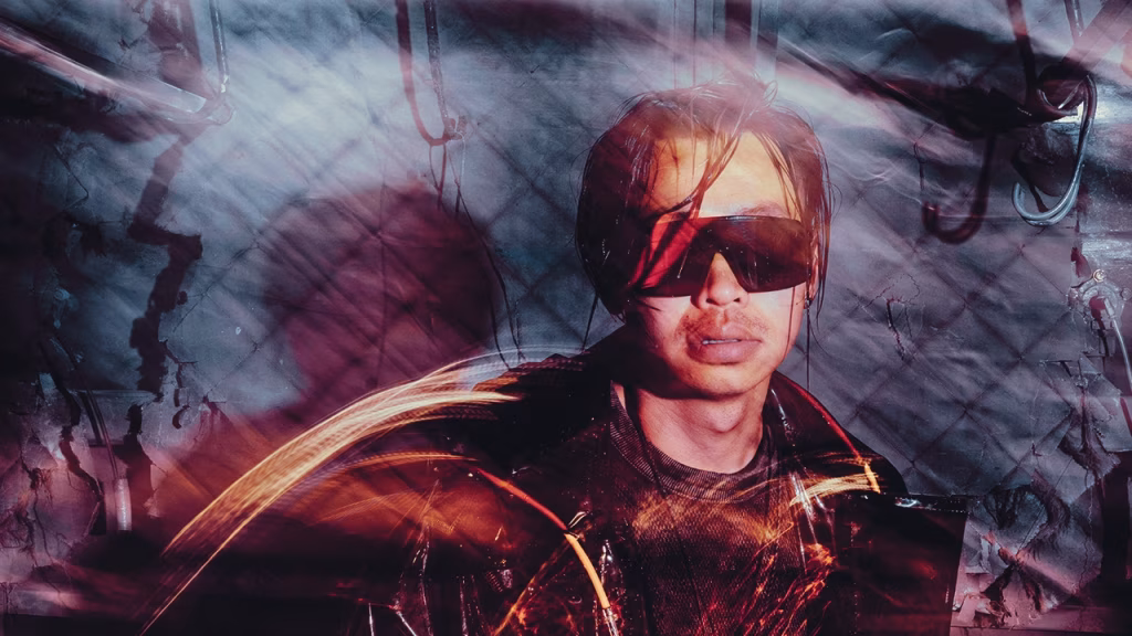 Close-up portrait of electronic music artist ZHU wearing sunglasses with red light trails, set against an industrial, textured background for the 'ZHU RIDES TO DREAMLAND' article. - midnightrebels.com