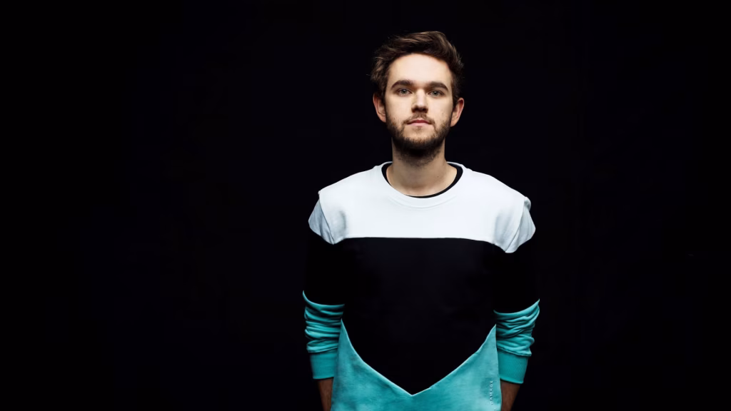 Zedd, the musician collaborating with Valorant, poses in a white, black, and teal color-blocked sweater against a black background. - midnightrebels.com