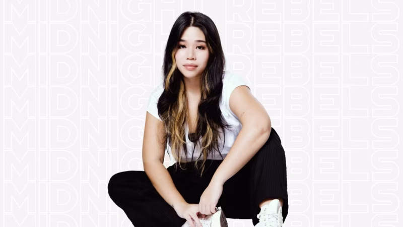 DJ A$trid, with long black hair and a blonde streak, sits cross-legged in a white t-shirt and dark pants against a light background with repeating text. - midnightrebels.com