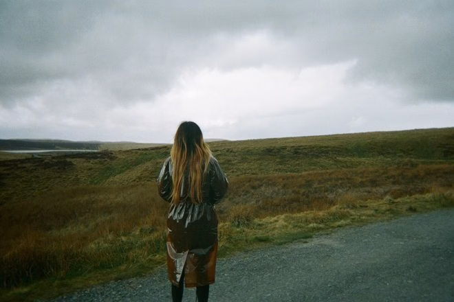 Flora Yin-Wong, facing away from the camera, stands on a gravel path overlooking a grassy landscape under a cloudy sky, representing the album's themes. - midnightrebels.com