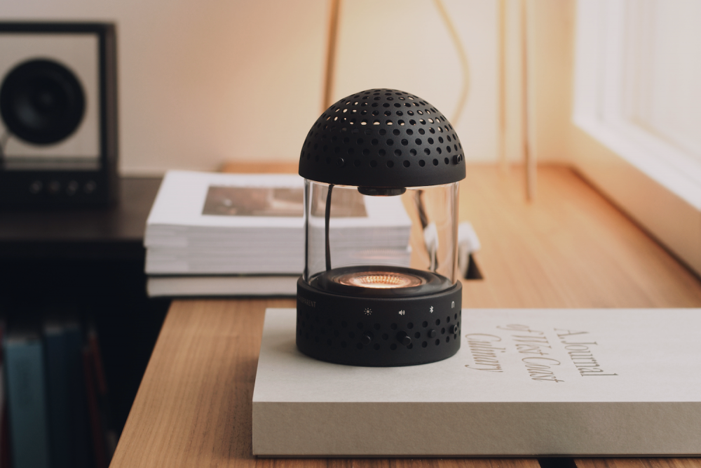 The TRANSPARENT portable light speaker, featuring a black perforated dome and clear cylindrical body with a warm light glow, sits atop a book on a wooden table in a bright, minimalist setting. - midnightrebels.com