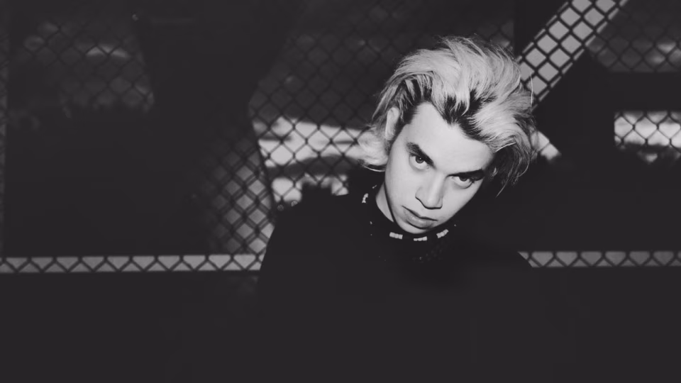 Black and white portrait of Ez Mil with dyed blonde hair, looking down at the camera in front of a chain-link fence. - midnightrebels.com