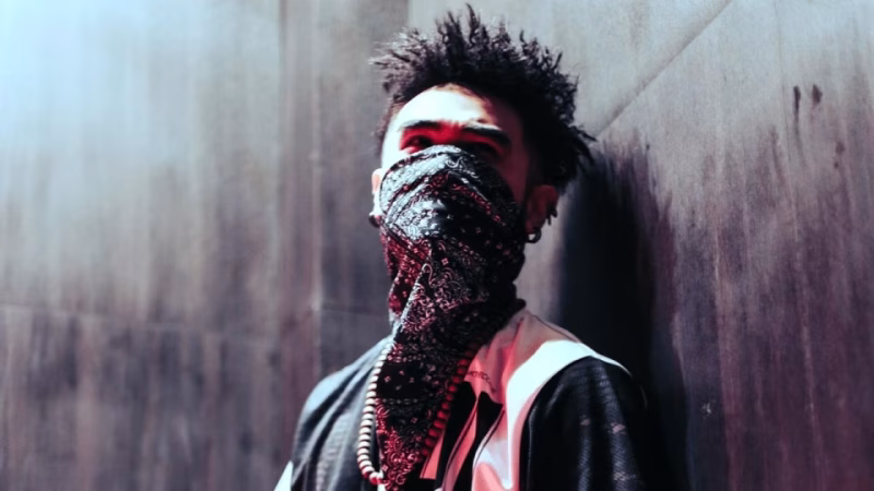 A person with dreadlocks and facial piercings stands against a textured wall, face obscured by a paisley bandana, representing the anonymity and 'stealing time' concept related to Bandit. - midnightrebels.com