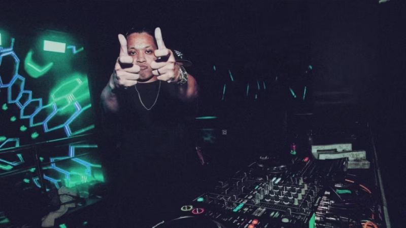 Kobeast, a DJ from Manila, strikes a pose with finger guns behind his DJ equipment in a dark club setting with green and blue geometric lighting. - midnightrebels.com