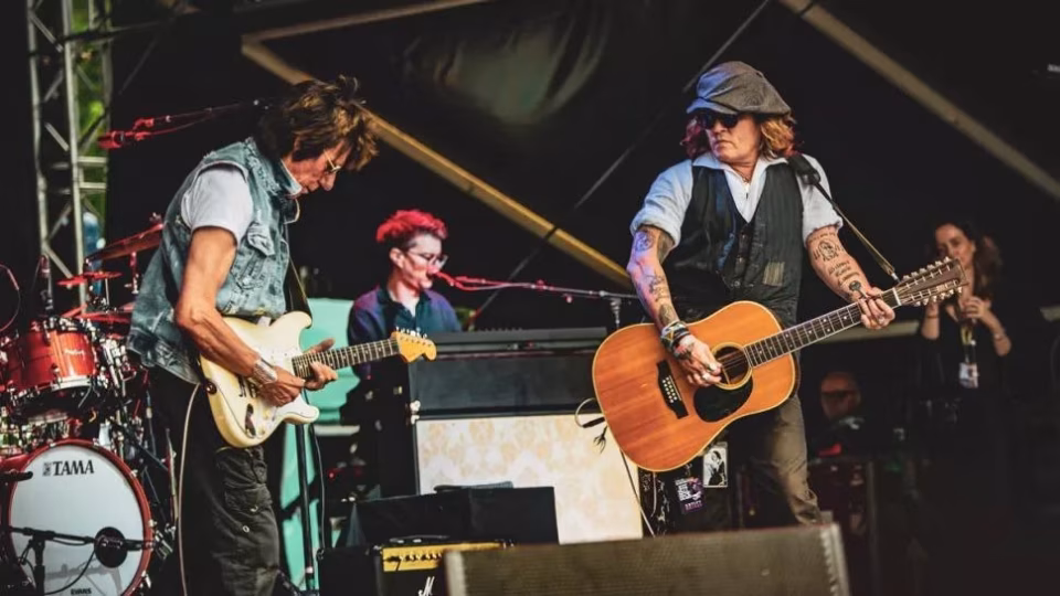 Johnny Depp, wearing a newsboy cap and tattoos visible on his arms, plays a 12-string acoustic guitar alongside guitarist Jeff Beck during a live performance. - midnightrebels.com
