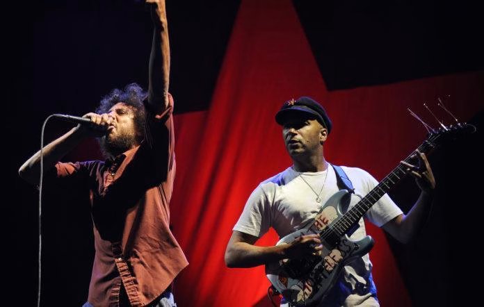 Rage Against the Machine's Zack de la Rocha passionately sings with a raised fist next to guitarist Tom Morello under a red star backdrop during a performance of "Born of a Broken Man." - midnightrebels.com