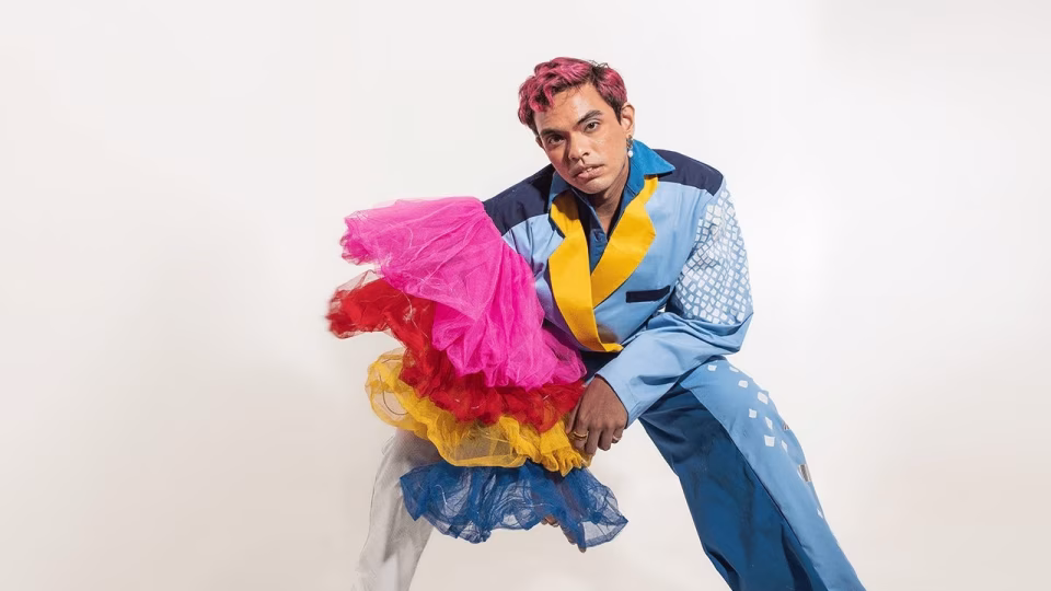 A person with pink hair in a blue patterned suit poses holding layered colorful tulle fabrics against a white background. - midnightrebels.com