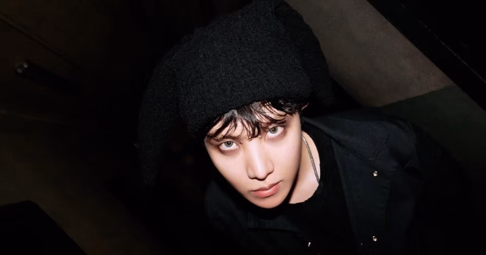 J-Hope of BTS, headlining Lollapalooza 2022, looks up with piercing eyes, wearing a black beanie and shirt against a dark background. - midnightrebels.com