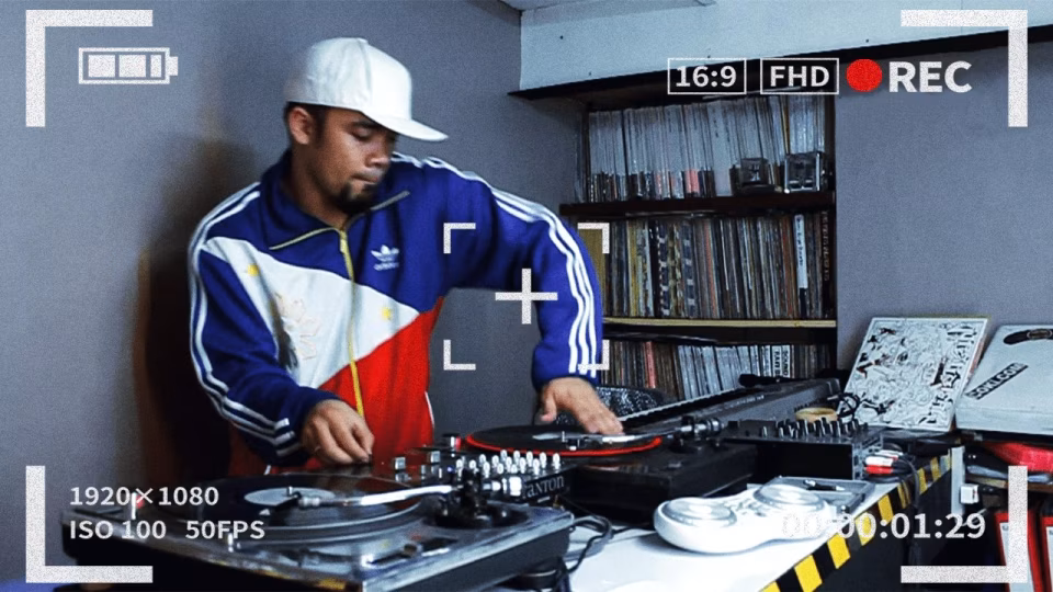 Through a camcorder-style viewfinder effect, DJ Coki is captured in action, mixing records on turntables with a record shelf in the background, for an article titled 'From the Deck to the Viewfinder There's No Stopping DJ Coki'. - midnightrebels.com
