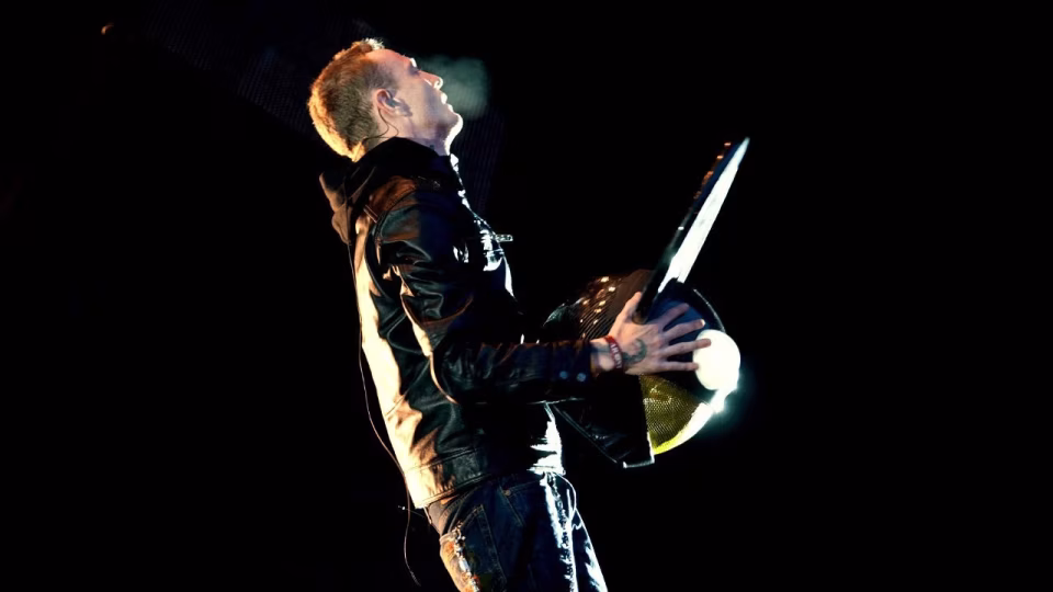 In a dimly lit, side-view shot, musician deadmau5, wearing a leather jacket and jeans, holds his iconic mouse-ear helmet while looking upwards, with visible breath. - midnightrebels.com