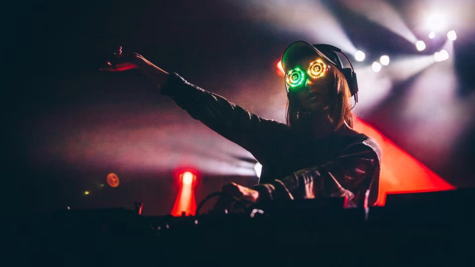 Rezz, wearing glowing goggles and headphones, performs as a DJ under stage lights. - midnightrebels.com