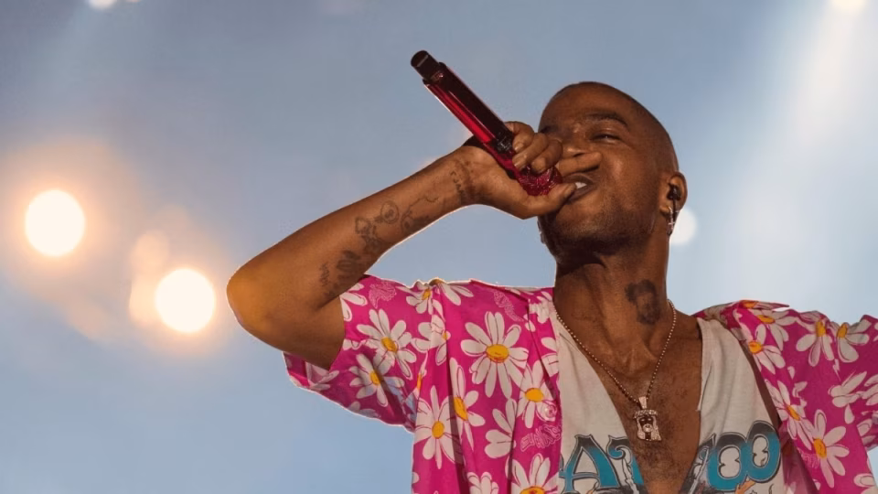 Kid Cudi, wearing a pink floral shirt and necklace, sings into a red microphone on stage, with tattoos visible on his arm and neck. - midnightrebels.com