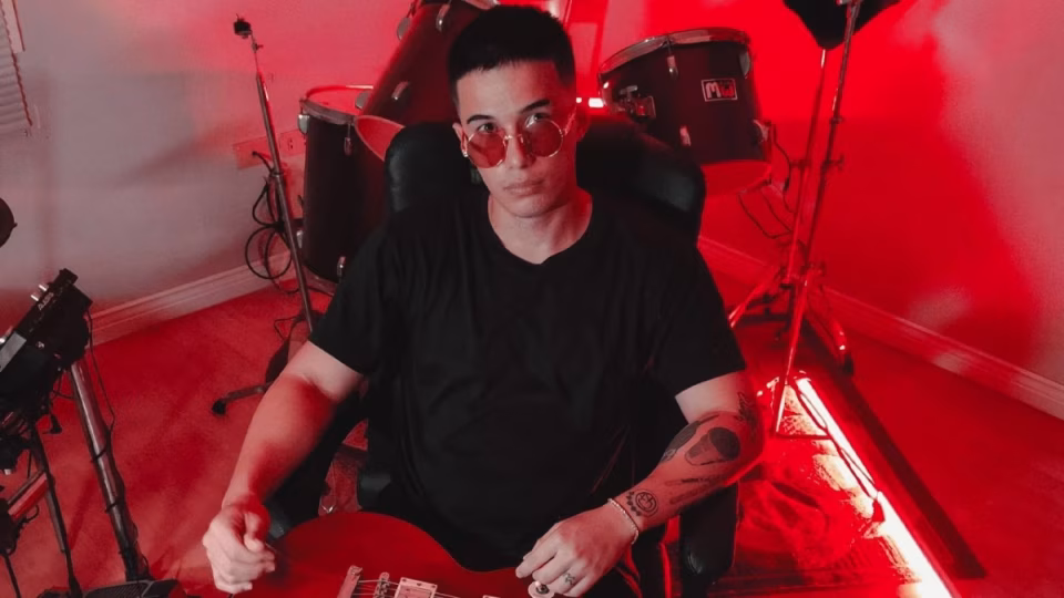 Mark Thompson, DJ and music producer, poses with a guitar under red lighting, surrounded by drum kit and music equipment. - midnightrebels.com