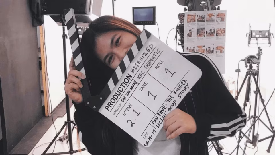 Sai, a filmmaker, smiles behind a clapperboard for "Bitesized" KFC thematic production, surrounded by film equipment. - midnightrebels.com