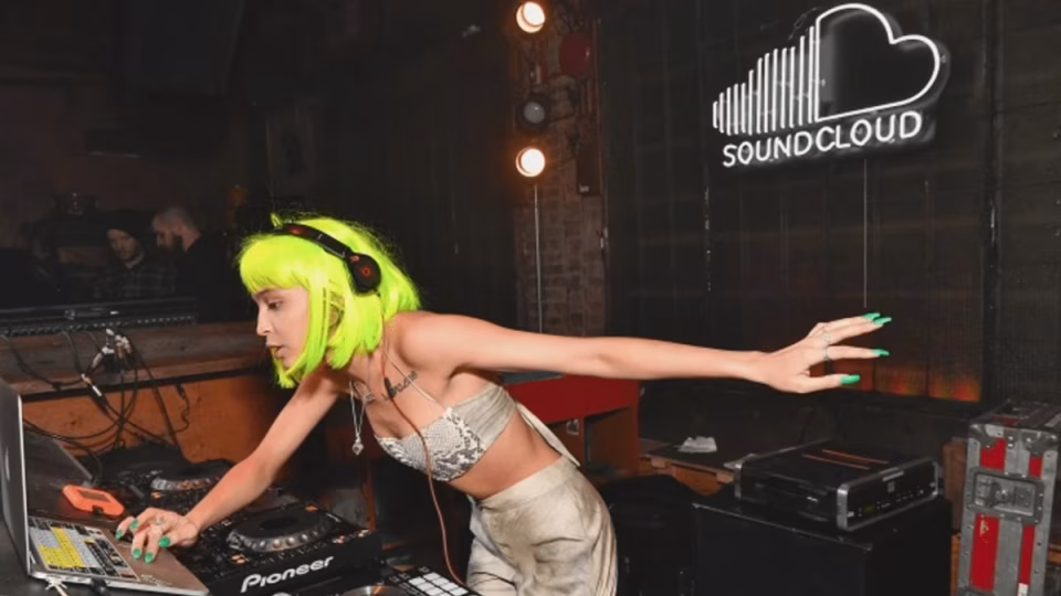 A female DJ with bright green hair and headphones is mixing music on Pioneer equipment in front of a neon SoundCloud logo. - midnightrebels.com