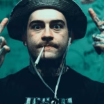 Getter, a heavily tattooed man with a cigarette in his mouth and wearing a bucket hat, holds up both middle fingers against a turquoise background. - midnightrebels.com