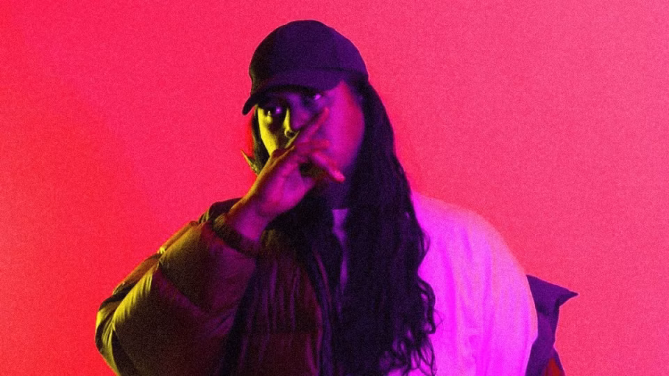 Montell2099, wearing a cap and jacket, poses against a vibrant pink backdrop with purple and yellow lighting, promoting his track "BLICKY". - midnightrebels.com