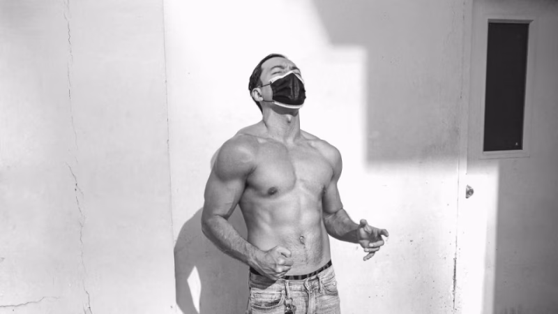 Black and white portrait of a muscular shirtless man, wearing a face mask, looking up in a moment of release, standing against a textured wall. - midnightrebels.com