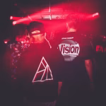 Under red rave lighting, the backs of three people are visible, one wearing a flamingo shirt and two in dark shirts printed with a triangle and the word "Vision" respectively, suggesting a vibrant nightlife scene in Asia. - midnightrebels.com