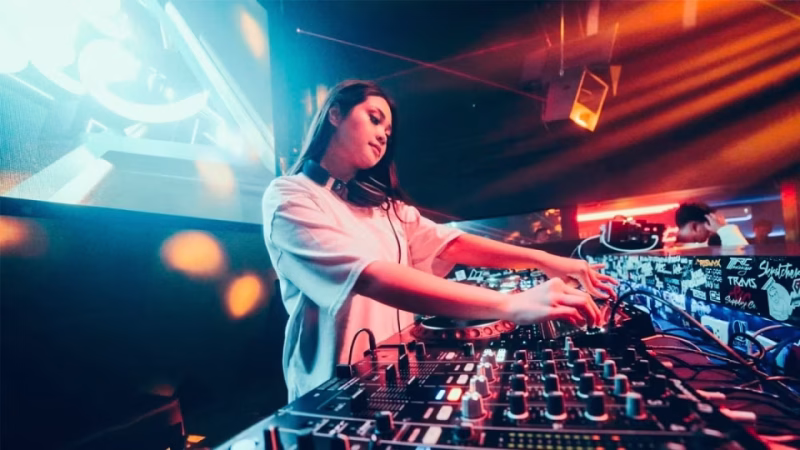 DJ Jade Dee is shown performing at a club, wearing headphones and a white shirt, focused on mixing music on her soundboard amidst colorful stage lighting. - midnightrebels.com