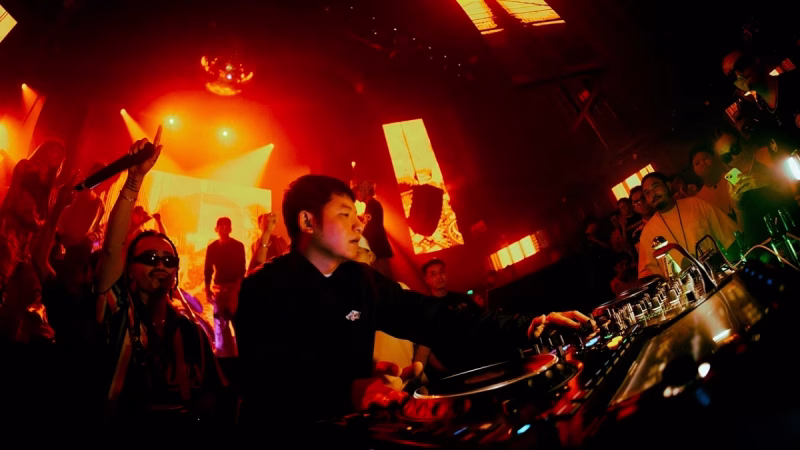Wide-angle shot of DJ Siangyoo performing live at a club, surrounded by a cheering crowd under red and orange stage lights, showcasing his digital DJing setup. - midnightrebels.com