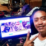 Close-up of DJ Love inside his recording studio, giving a peace sign in front of a computer monitor displaying a vibrant Budots music video. Cables and equipment surround the setup, giving a glimpse into the creative process. - midnightrebels.com