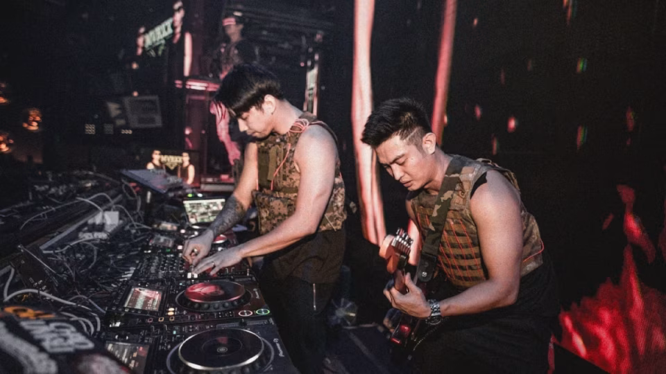 Two Filipino DJs in camo vests perform with turntables and a guitar at a nightclub, illustrating the evolving music scene in the Philippines. - midnightrebels.com