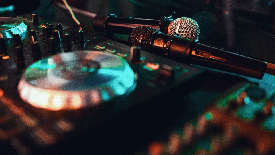 A close-up, low-lit shot of a DJ controller and two microphones represents the DJing setup. - midnightrebels.com