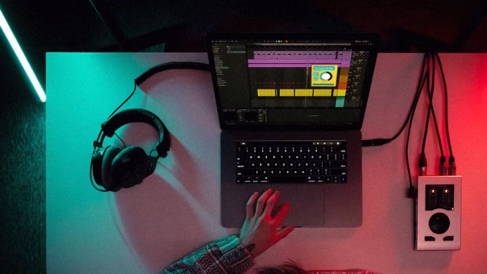 Overhead shot of a person working on music production software on a laptop, with headphones and an audio interface under red and teal lighting. - midnightrebels.com