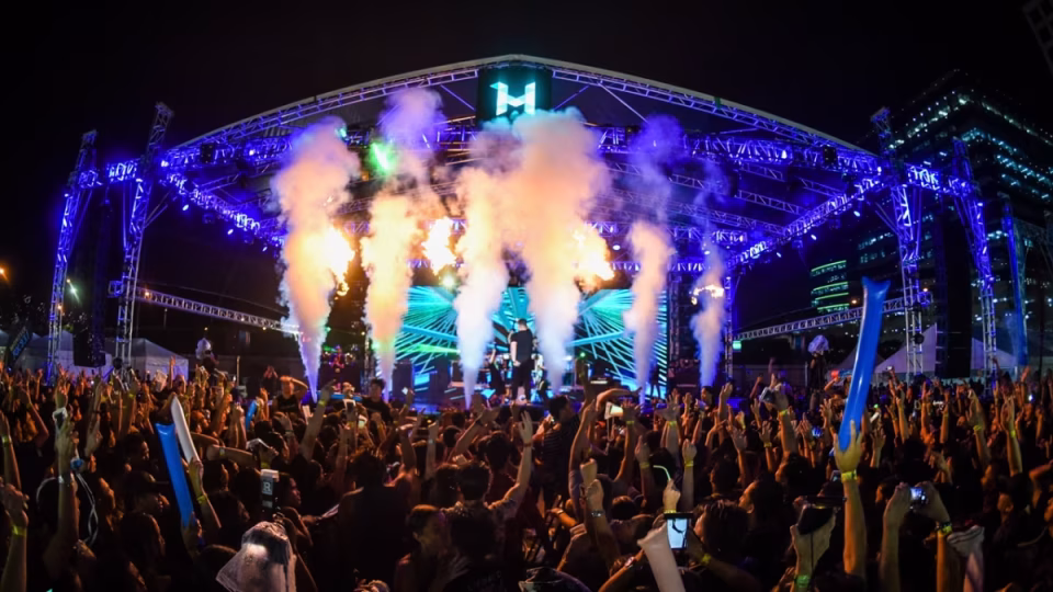 A large crowd with hands raised enjoys the Hydro Manila rave, featuring a brightly lit stage with performers, pyrotechnics, and smoke effects. - midnightrebels.com