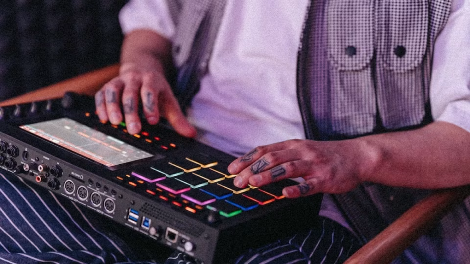 A DJ with hand tattoos uses a modern beat machine with colorful, lit-up pads to create music. - midnightrebels.com
