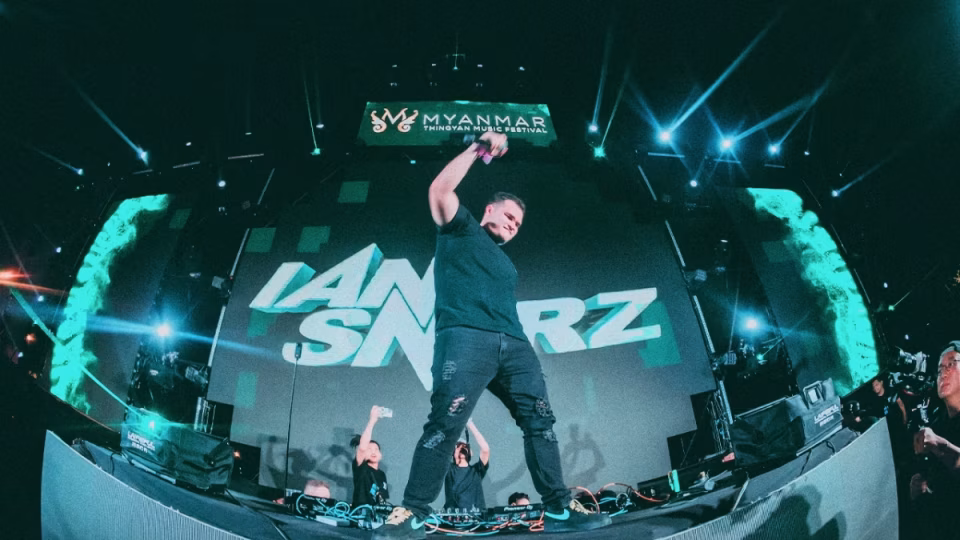 Ian Sndrz commands the stage at the Myanmar Thingyan Music Festival, bathed in teal lights and projected visuals, energizing the crowd with microphone in hand. - midnightrebels.com