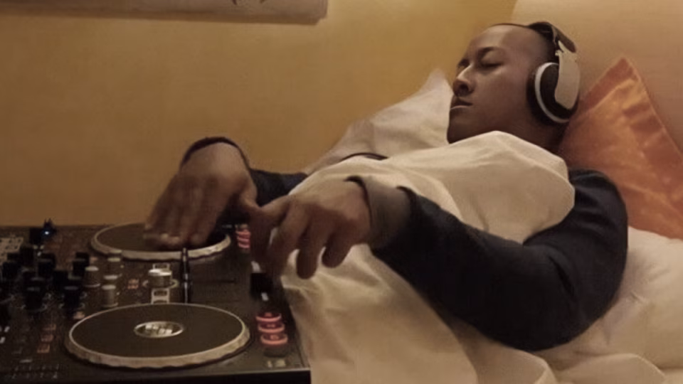 A tired DJ wearing headphones lies in bed with a blanket, seemingly asleep while still resting a hand on a set of turntables. - midnightrebels.com