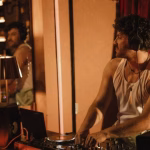 Professional male DJ wearing headphones and a white tank top, mixing tracks on a turntable in a dimly lit venue with red curtains, reflecting on the lifestyle and challenges of maintaining relationships. - midnightrebels.com