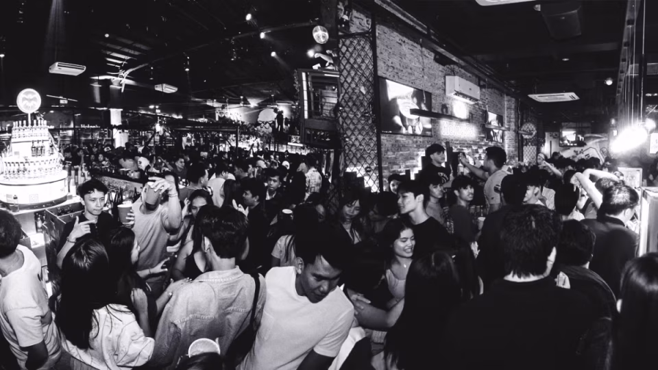 Black and white image of a densely packed crowd at a nightlife venue, likely Sentral or Proof in Cebu, Philippines, reflecting the vibrant energy of the city's nightlife scene. People are seen socializing and enjoying drinks, contributing to a lively atmosphere. The bar is fully stocked with bottles, and neon signs decorate the exposed brick walls. - midnightrebels.com