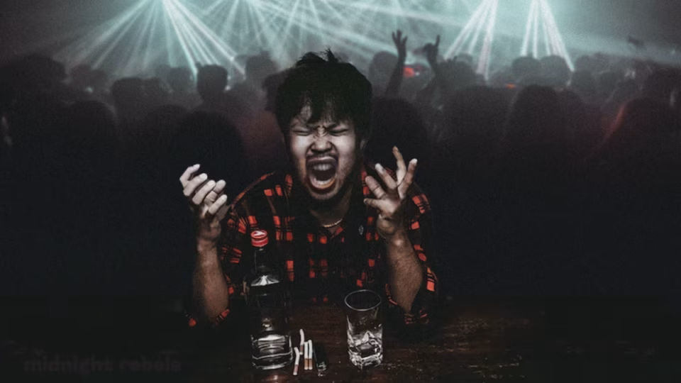 Frustrated clubber yelling at a table with alcohol and cigarettes in a crowded nightclub. Image conveys the feeling of being burned out on overplayed club hits. - midnightrebels.com