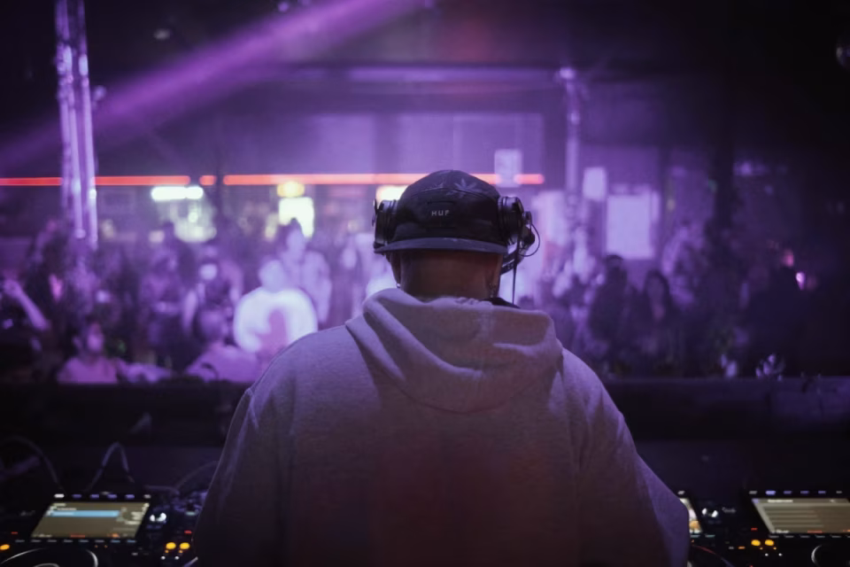 Back view of a DJ in a purple-lit club, standing behind mixing equipment with a blurred crowd in the background, representing the debate between algorithmic DJing and artistic expression. - midnightrebels.com