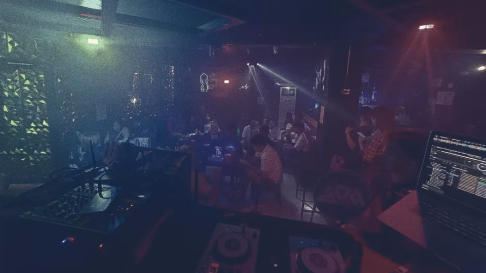 View from a DJ booth in a crowded nightclub, emphasizing the tension and potential for conflict over song requests. Equipment including a laptop with DJ software and turntables are visible. The crowd is dimly lit and engaged in socializing. - midnightrebels.com