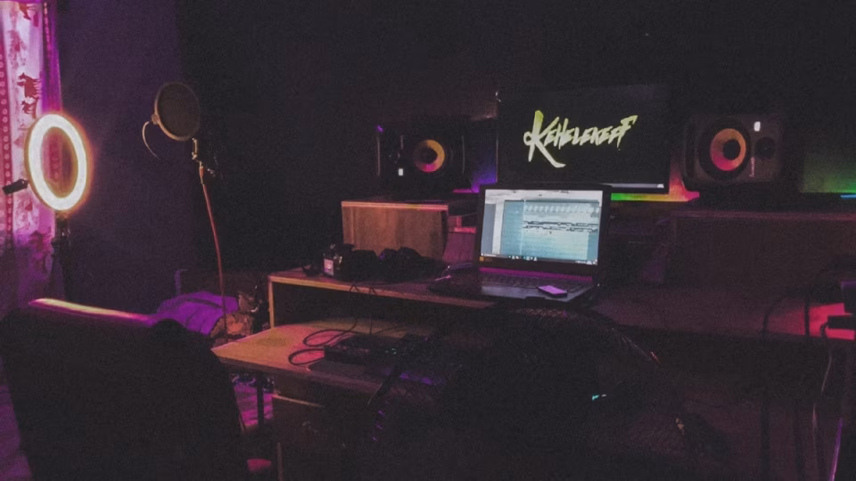 A dimly lit home music production studio setup with a laptop running FL Studio, studio monitors, a microphone with pop filter, and a ring light, questioning the necessity of high-end equipment for creating great music. - midnightrebels.com
