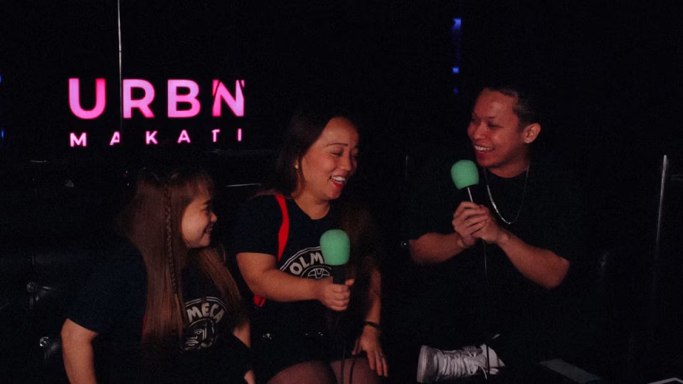 A group of three individuals are gathered at URBN Makati, laughing and talking into green microphones. The pink URBN Makati sign is visible in the background, highlighting the venue's importance in fostering community and sparking social conversations. - midnightrebels.com