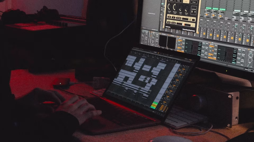 Close-up of a Filipino DJ-Producer working on music production software (Ableton Live) on a laptop and monitor in a dimly lit studio. Hands are visible on the keyboard, suggesting intense focus and dedication. The image represents the hard work and technical skills required for success in the competitive electronic music scene. This image is for an article about Filipino DJ-Producers and the challenges they face in achieving breakthrough success. - midnightrebels.com