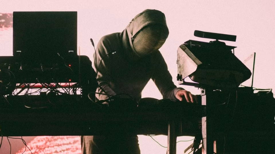 Silhouetted figure in a hoodie and mesh mask performs techno music behind a complex DJ setup with screens, cables, and equipment, highlighting the anonymity and mystery sometimes associated with new-wave techno artists and sparking questions about authenticity in the age of clout. - midnightrebels.com