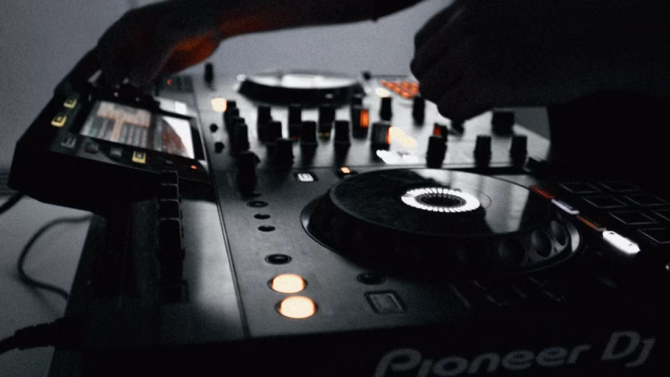 Close-up shot of a Pioneer DJ mixer being operated in a dimly lit club setting. The hands of the DJ are visible, adjusting knobs and faders. This image illustrates the tools of the trade as DJs navigate new venue policies requiring them to bring guests to secure their set. - midnightrebels.com
