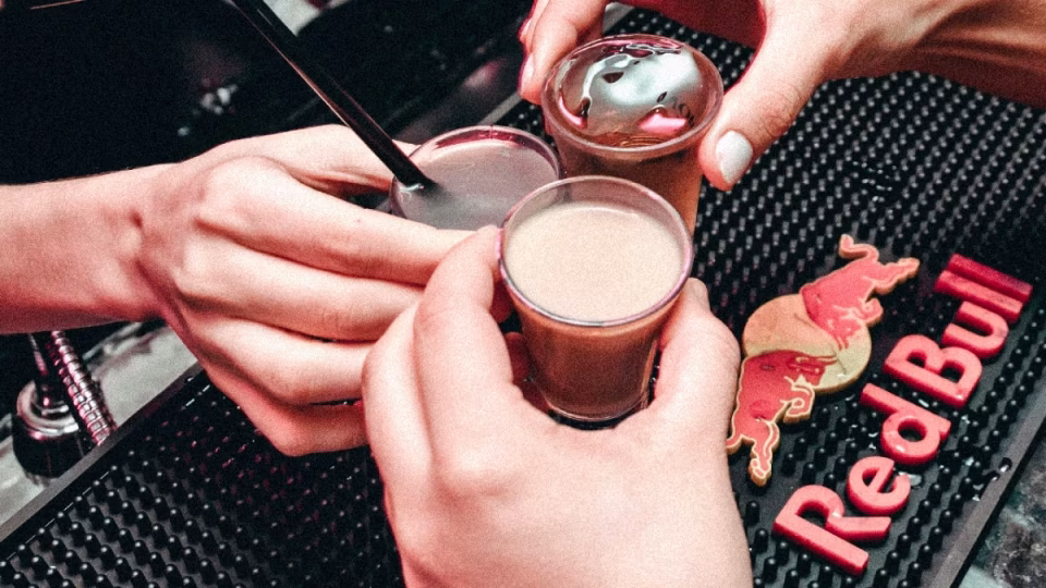 Close-up of three hands holding shot glasses filled with colorful cocktails on a bar counter with a Red Bull logo, representing potential factors influencing bar sales including drink choices and brand appeal, relevant to discussions about DJs and bar revenue. - midnightrebels.com
