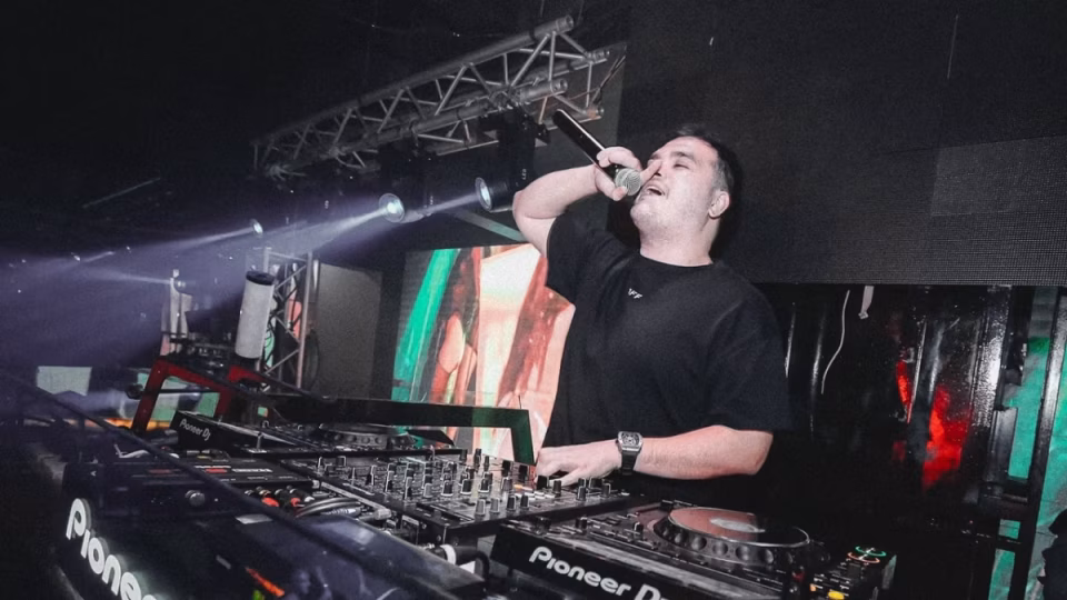 Ian Sndrz performing live, creating the magic behind the groundbreaking global Christmas mashup pack. He is singing into the microphone while working with the DJ mixing board. A large screen and stage lighting are visible in the background. - midnightrebels.com