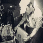 Black and white image of a DJ selecting a vinyl record from a crate at a live club event. The scene is packed with attendees and photographers, emphasizing the performative and curated nature of modern DJing. - midnightrebels.com
