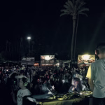 DJ performing a silent disco set at a crowded night market, illuminated by food stalls and overhead lighting. Attendees are wearing glowing wireless headphones. - midnightrebels.com