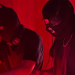 Under red lighting, two DJs wearing masks and headphones play bass music in Cebu, with a disco ball and a glowing sign visible. - midnightrebels.com